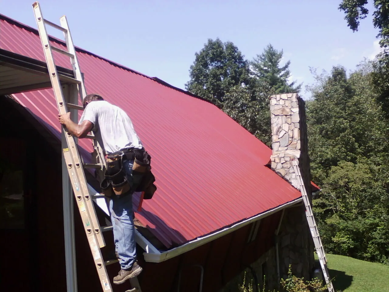 Roofer on a ladder beside a red metal roof and stone chimney — Soffit & Fascia Repair by Roofing Brothers in Toney